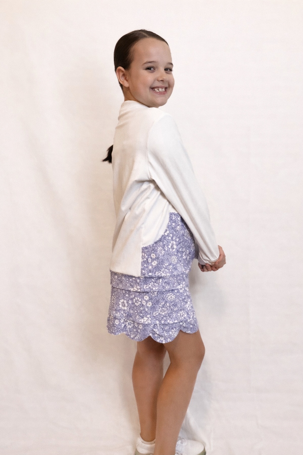 Side view of a young girl wearing a white long sleeve top with large floral pocket and floral, scalloped hem skirt on a plain background