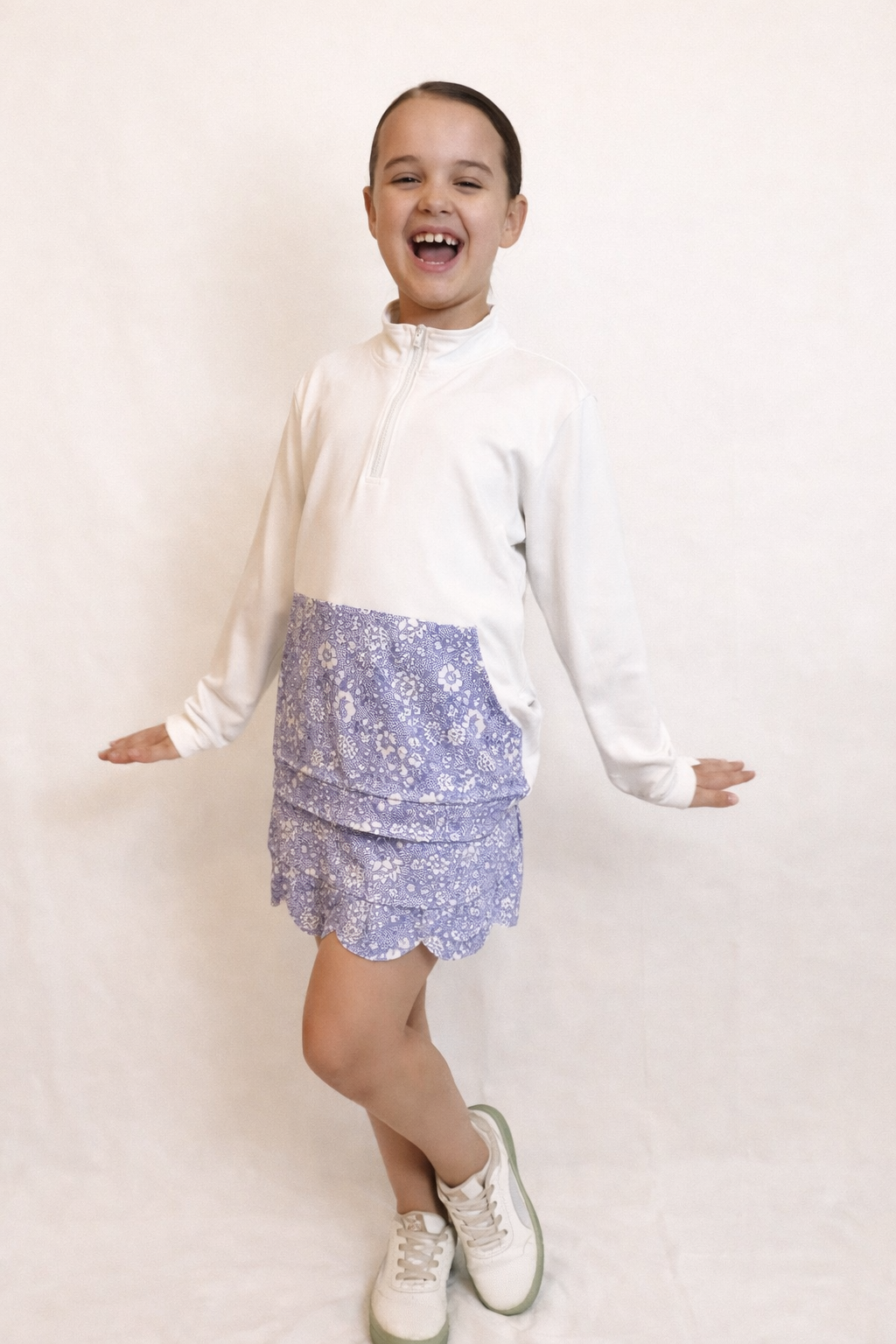 Young girl wearing a white long sleeve half zip top and floral skirt set on a plain background