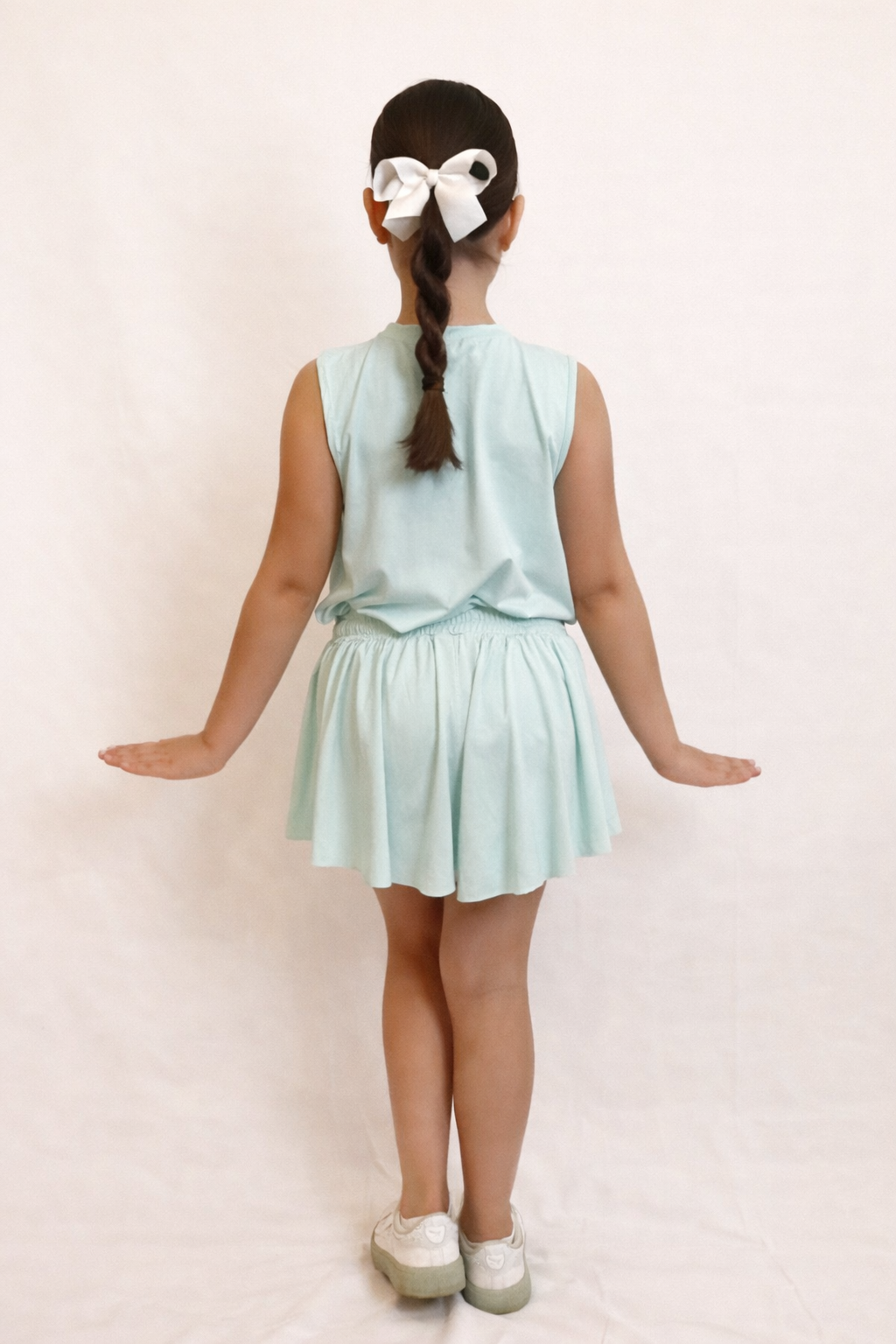 Back view of a young girl wearing a mint green sleeveless top and flowy skort bottom against a plain background