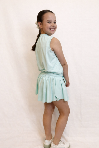 Side view of a young girl wearing a mint green sleeveless top and flowy skort with side slits against a plain background
