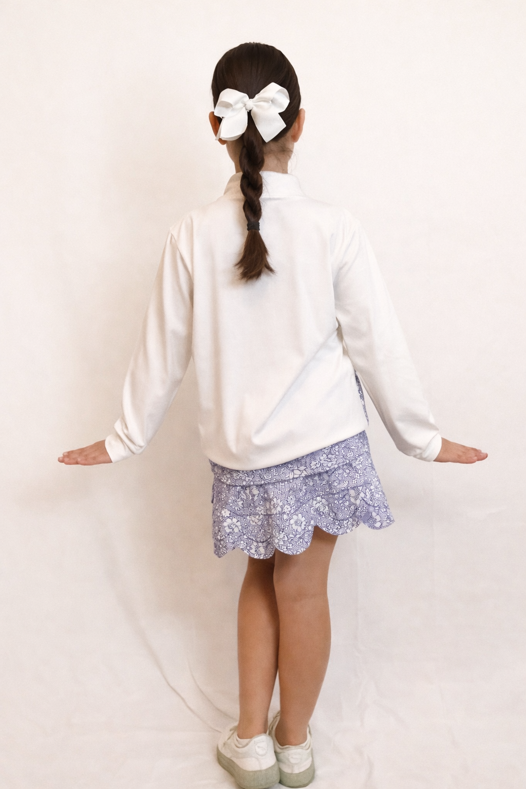 Back of young girl wearing a white long sleeve top with floral bottom band and floral skirt with scalloped hem on a plain background