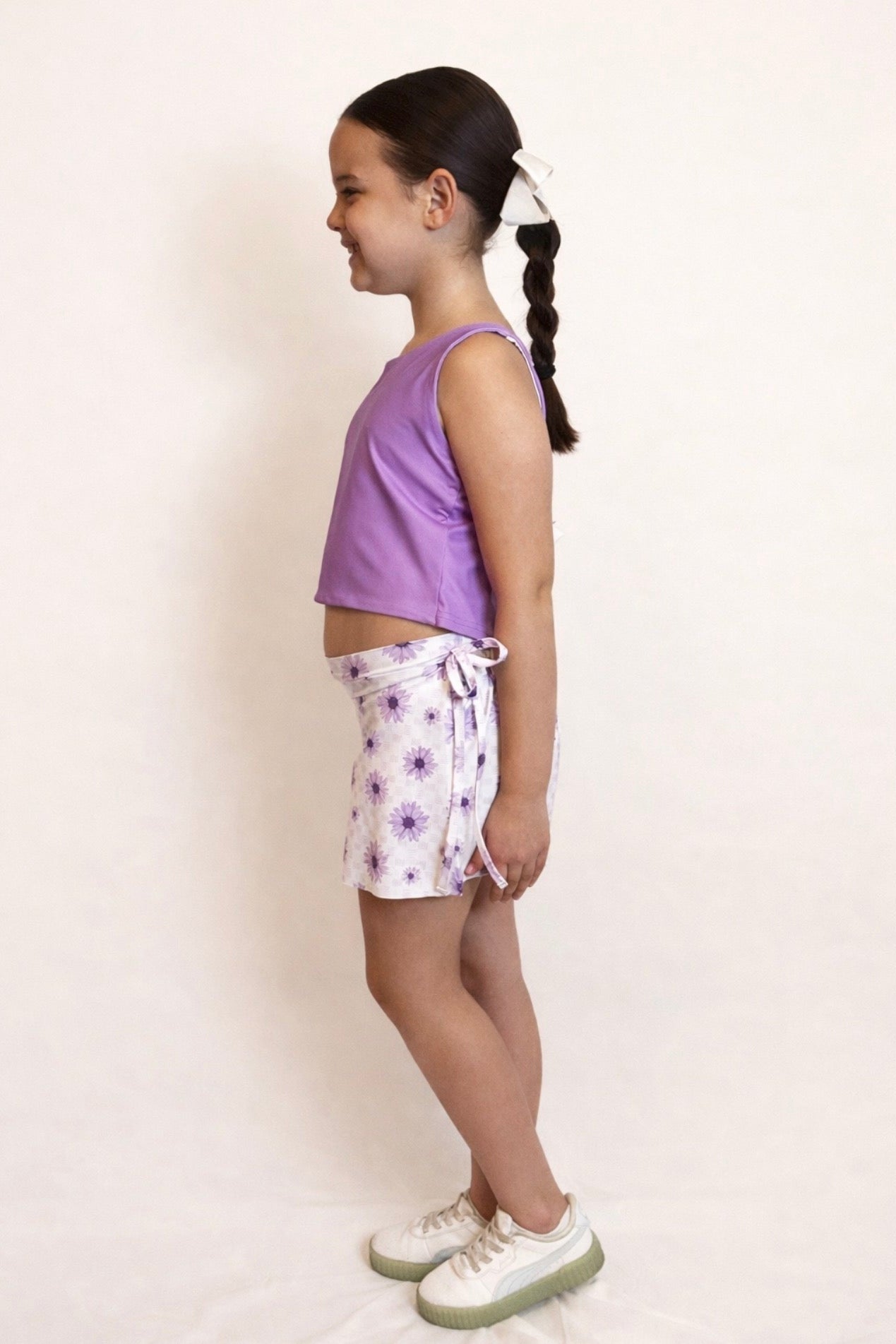 Side view of a young girl wearing a purple crop top and floral patterned shorts with faux bow tie on a white background