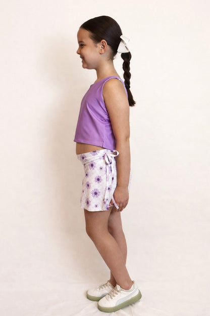 Side view of a young girl wearing a purple crop top and floral patterned shorts with faux bow tie on a white background